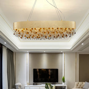 The Avani Crystal Dining Room Chandelier by Morsale.com is a modern luxury chandelier with a sleek, circular design. It features an arrangement of amber and clear crystals and gold accents hanging below a wide, gold band. The chandelier is suspended from the ceiling by thin wires, set against a plain gray background.