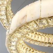 Close-up of a luxurious, modern ceiling chandelier featuring two interlocking, circular light fixtures. The lights are adorned with square-cut crystal accents and white marble-like panels, creating an elegant design. This Morsale.com Natural Marble & Crystal Modern Ceiling Light Fixture emits a warm, radiant glow.