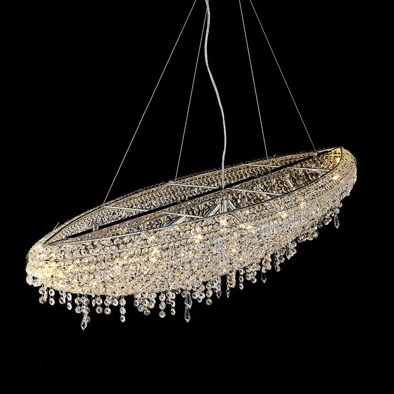 The Alleri Crystal Chandelier for Dining Room from Morsale.com is an elegant, elongated chandelier with intricate detailing and handmade crystal droplets. Suspended against a black background, its oval design features multiple rows of crystals, illuminated by E14 LED bulbs that reflect a warm light, enhancing its sophisticated appearance.