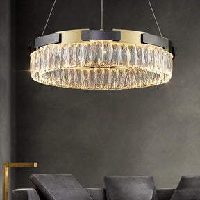 A sophisticated Siena Contemporary Crystal Light Fixture from Morsale.com with a luxurious finish and detailed clear crystal elements hangs above a modern, dark grey couch. The chandelier features a gold and black frame, adding an elegant touch to the dimly lit room with dark grey walls. A brass side table is partially visible.