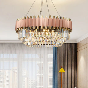 A Gio Rose Gold Modern Chandelier from Morsale.com with a geometric design featuring gold and pink rectangular elements hangs from the ceiling in a well-lit room. Handcrafted crystals add elegance, while sheer curtains, a gray sofa, and a yellow floor lamp complete the look.