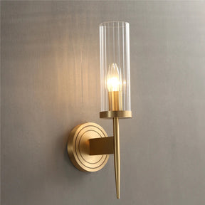 The Palermo Postmodern Copper Wall Sconce by Morsale, featuring a circular copper base and a tall, cylindrical glass shade, casts warm light onto the textured gray wall, highlighting its sleek, minimalist design. This stylish lighting piece adds an elegant touch to any room decor.