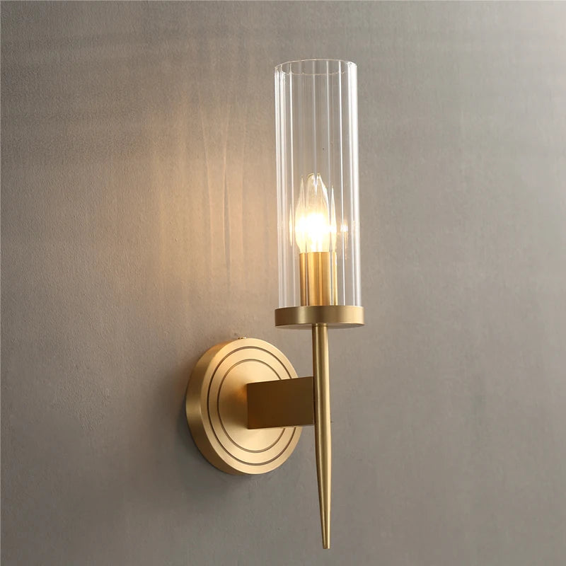 The Palermo Postmodern Copper Wall Sconce by Morsale, featuring a circular copper base and a tall, cylindrical glass shade, casts warm light onto the textured gray wall, highlighting its sleek, minimalist design. This stylish lighting piece adds an elegant touch to any room decor.