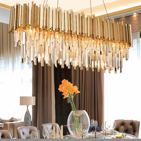 The luxurious dining room showcases a modern Gio Crystal Dining Room Chandelier by Morsale.com, with gold and glass cylindrical elements, featuring LED bulbs, hanging over a table set for a meal. At the center of the table is an elegant glass vase with orange flowers, surrounded by wine glasses and upholstered chairs.