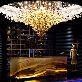 A luxurious interior showcases a large Piovere Multi Pendant Chandelier by Morsale.com with a crystal shade above an onyx reception desk. A warrior statue and decorative wall elements enhance the opulent atmosphere, highlighted by warm luxury lighting.