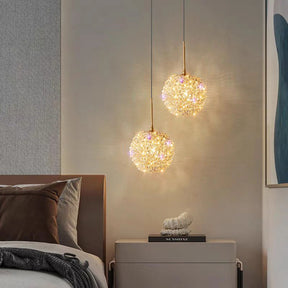 A cozy bedroom features El Sol Crystal Pendant Light Fixtures by Morsale.com above a nightstand, providing energy-efficient illumination. The bed has brown and white pillows, a sculpture on the nightstand, and minimalist wall art is visible in the warm glow.