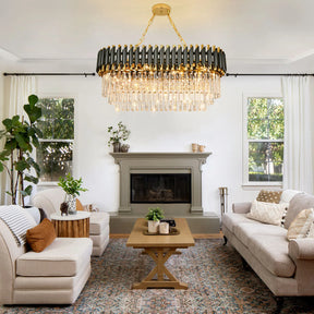 A stylish living room featuring a large, modern **Verano Crystal Dining Room Light Fixture** by **Morsale.com** with dimmable lights hanging above a wooden coffee table, flanked by two beige sofas with patterned cushions. The room has a decorative fireplace, lush potted plants, and large windows with white curtains enhancing the natural light and contemporary design.
