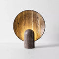 The Morsale Natural Travertine Table Lamp features a cylindrical base with a large circular backplate. Crafted from natural travertine, it emits a warm, golden glow. This vintage design combines minimalist aesthetics with adjustable brightness, set against a plain white background for a timeless look.