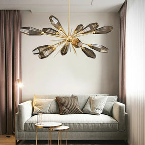 A modern living room with a light gray sofa adorned with pillows, a round side table, and a floor lamp. A striking Morelli Sputnik Chandelier from Morsale.com with multiple black-shaded bulbs hangs from the ceiling. Large windows with sheer curtains let in natural light.