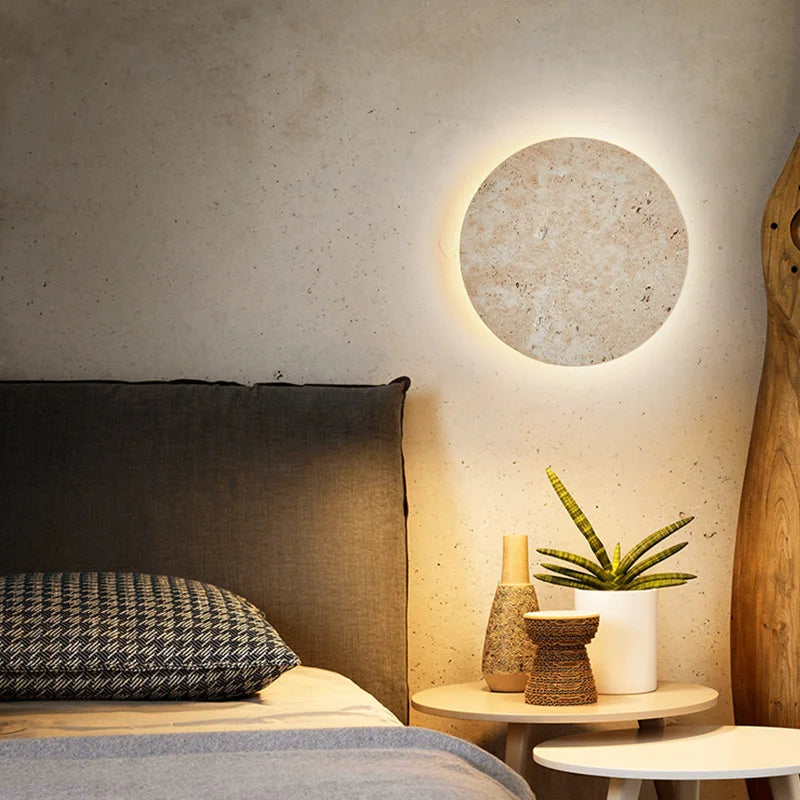 A cozy bedroom featuring a modern Morsale.com Natural Travertine Wall Sconce emitting a soft glow, a dark headboard, and a bedside table. On the table are a small plant in a white pot and two textured brown vases, contributing to the warm and inviting ambiance.