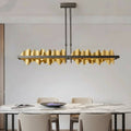 A modern dining room features a white marble table with beige chairs and the Rivaro Brass Linear Chandelier by Morsale.com above. An open book and a decorative bowl adorn the table.