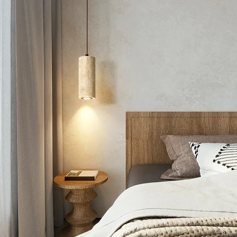 A cozy bedroom corner with a wooden headboard, round wood side table with a book, gray curtains, layered neutral bedding, decorative pillows, and warm natural stone lighting from the Brio Travertine Pendant Light by Morsale.com.