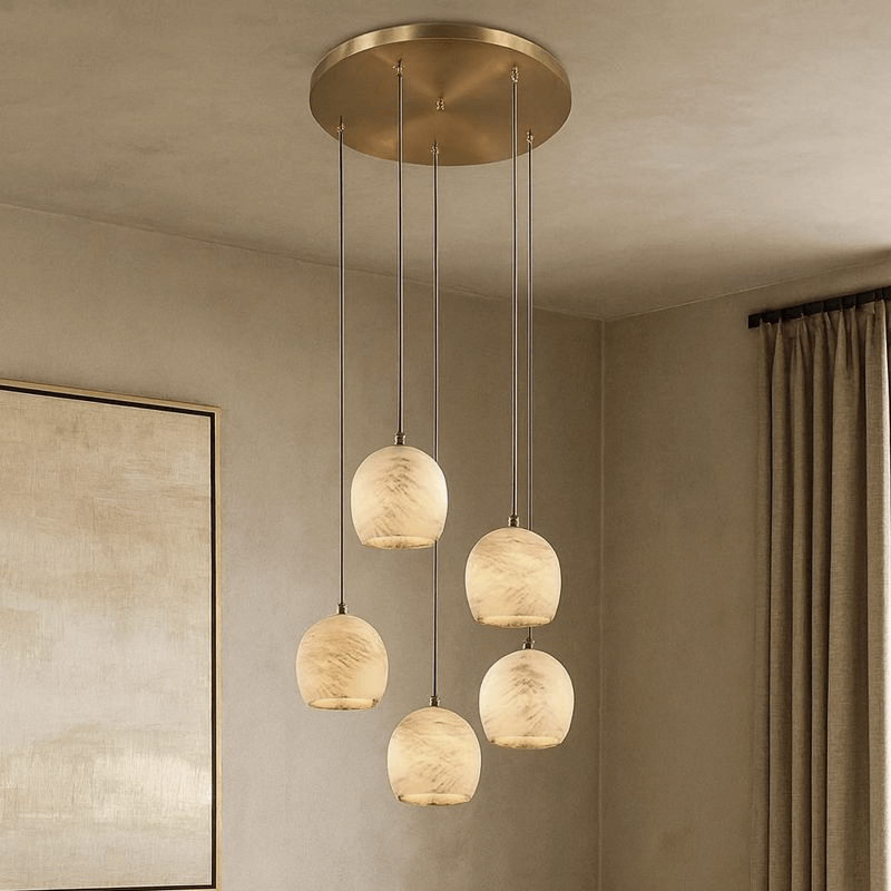 The Auris 5-Light Marble Chandelier from Morsale.com features five creamy marbled glass pendants suspended at different heights from a brass circular mount with adjustable cords, perfect for neutral rooms with framed art and beige curtains.
