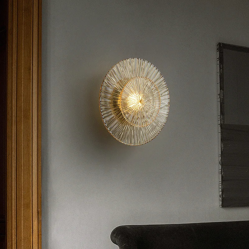 A Modern Glass Wall Sconce by Morsale.com with an intricate, gold wire design is mounted on a grey wall. This handmade fixture illuminates with a warm light. A wooden door frame is partially visible on the left, and the back of a dark-colored sofa is at the bottom right.