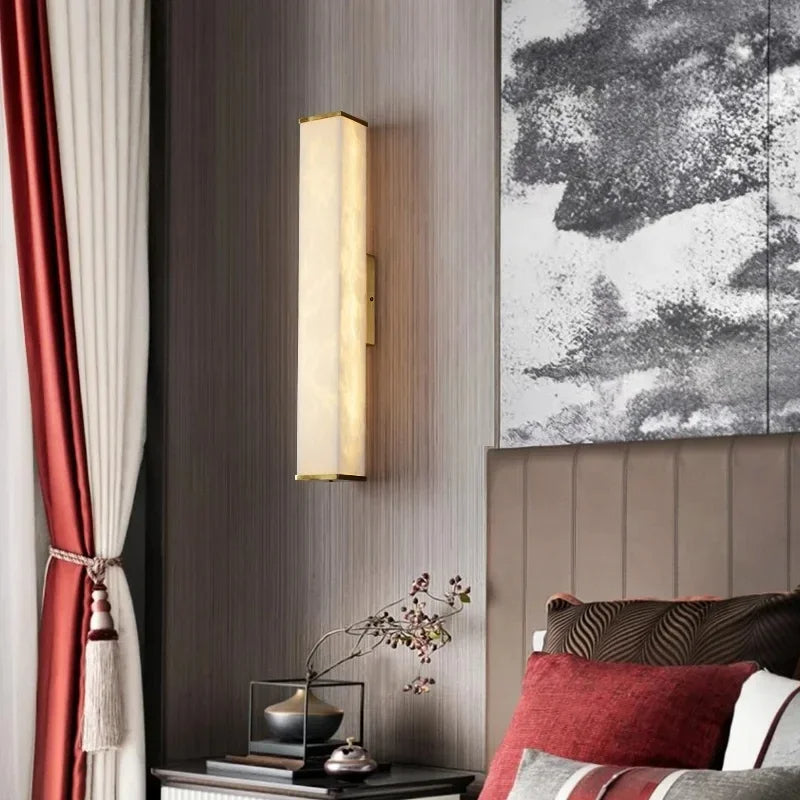A modern bedroom features a Morsale.com Natural Marble Indoor Wall Sconce Light with a rectangular design, casting warm light on a textured wall. The bedside table holds a minimalist arrangement with a small vase and decorative items. Red and white curtains frame the window, complementing the bed's color scheme.