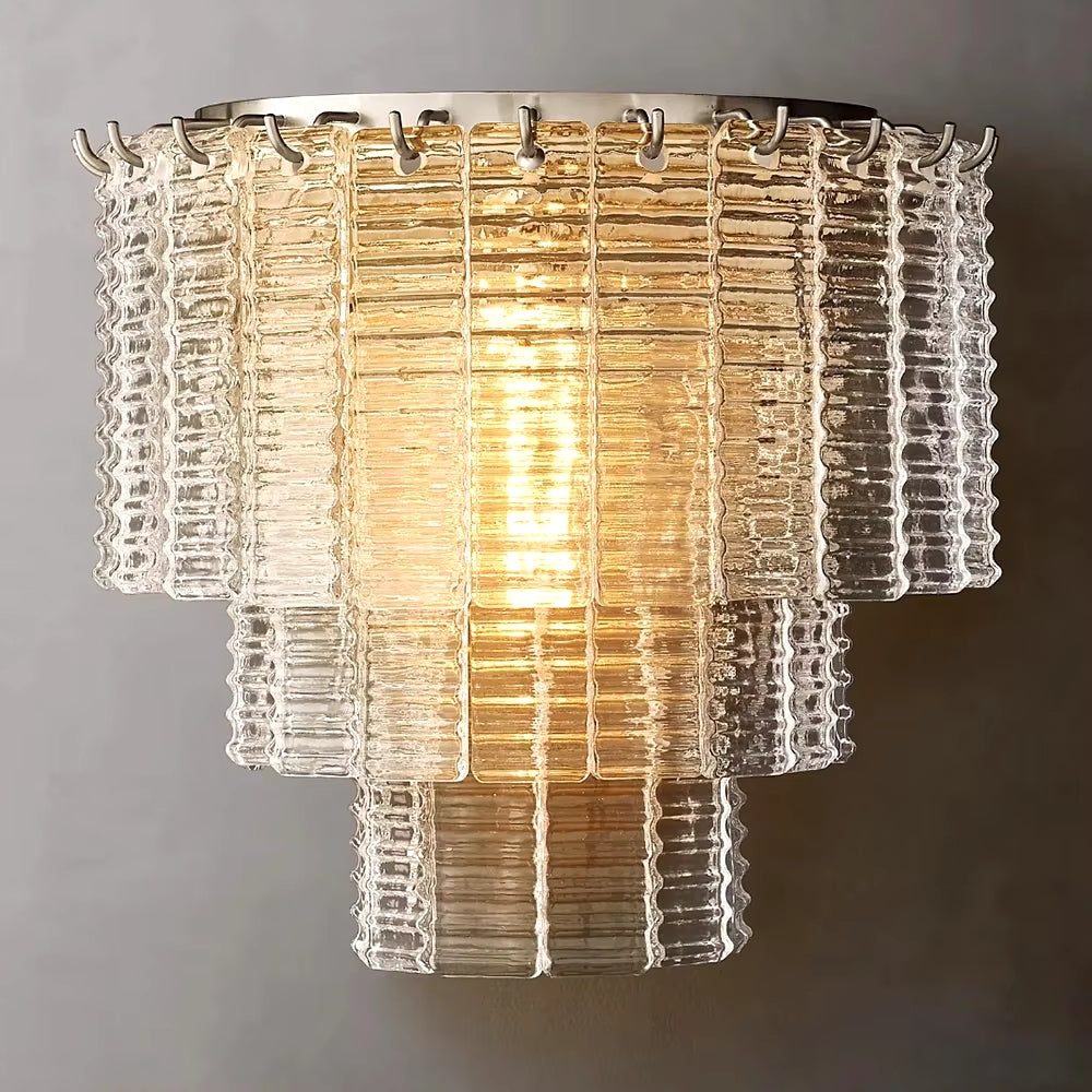 The Palermo Clear Crystal Wall Sconce, by Chandelier Style, showcases a tiered design with stacked, textured glass panels. The clear glass shade creates a layered effect around the warm light and is enhanced by crystal accents for a glowing appearance against a neutral background.