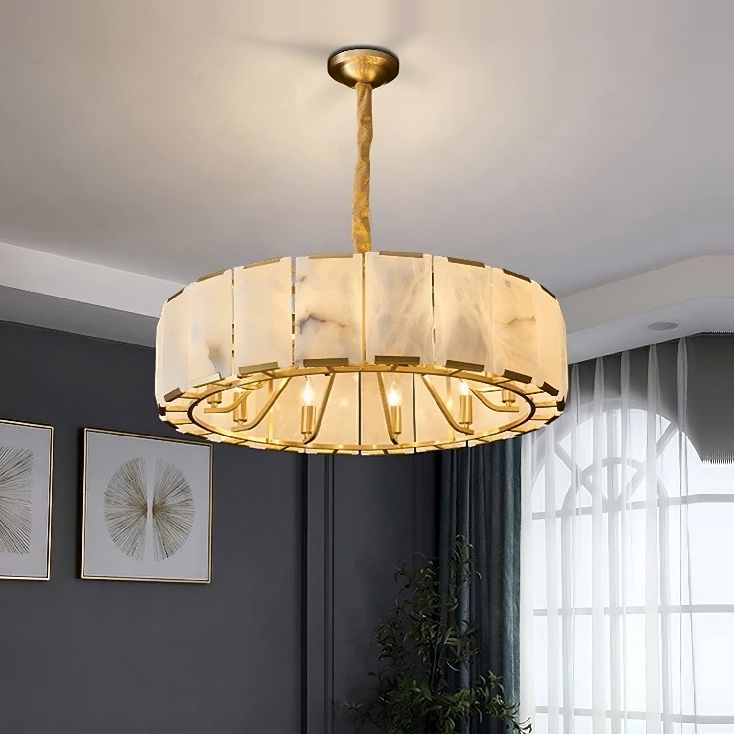 A modern Natural Marble Ceiling Chandelier by Morsale.com with a cylindrical, semi-translucent white and gold frame hangs from the ceiling. Below, the room features dark walls, framed geometric artwork, a large window with sheer white curtains, and a potted plant in the corner. The G9 LED bulbs add an elegant glow to the space.