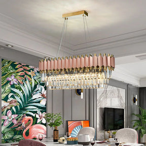 A modern living room adorned with a large, rectangular crystal chandelier featuring vertical prisms and gold accents. The room boasts vibrant flamingo-themed wall art, a gray sofa, a green plant, and a small wooden-topped table, all illuminated by the Gio 39" Rose Gold Dining Room Chandelier from Morsale.com near the TV mounted on the gray wall.