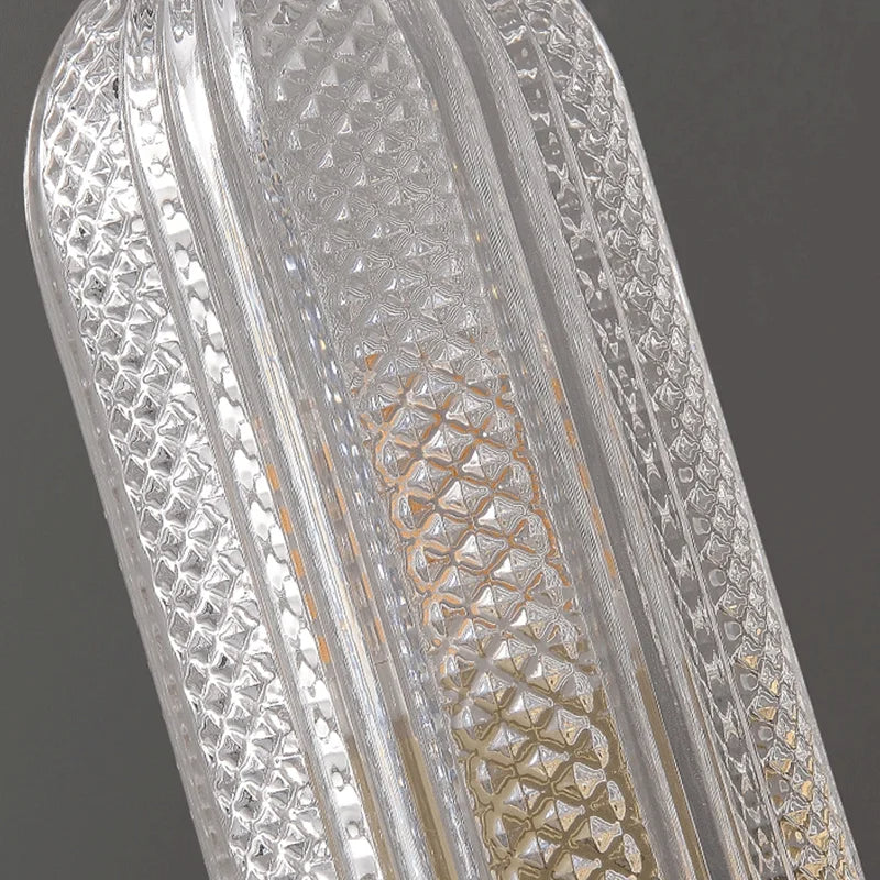 Close-up of a textured, transparent light fixture with an elongated shape. The intricate, geometric patterns on the outer surface create a shimmering effect, with a visible central element inside. This Morsale Palermo 12" Brass Wall Light Sconce offers stylish design and warm lighting against a neutral gray background.