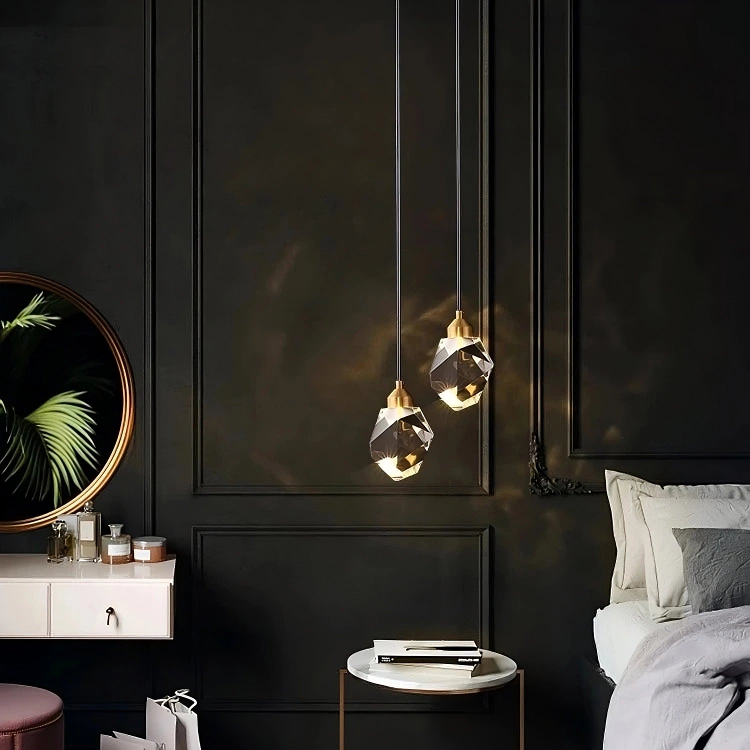 In a contemporary bedroom with dark walls, two Diamante Crystal Pendant Lights by Morsale hang elegantly above a round white side table. A large round mirror reflects the greenery of plant leaves, positioned next to a neatly made bed adorned with white bedding on the right.