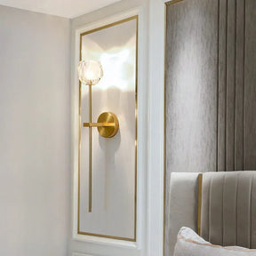 A Clear Crystal & Brass Wall Light Fixture by Morsale.com is mounted on a white and gold-trimmed wall panel. With its gold finish and clear crystal globe shade, this elegant modern sconce provides ambient lighting next to a bed featuring a gray, vertically paneled headboard. Part of the bed and a fluffy pillow are visible in the setup.