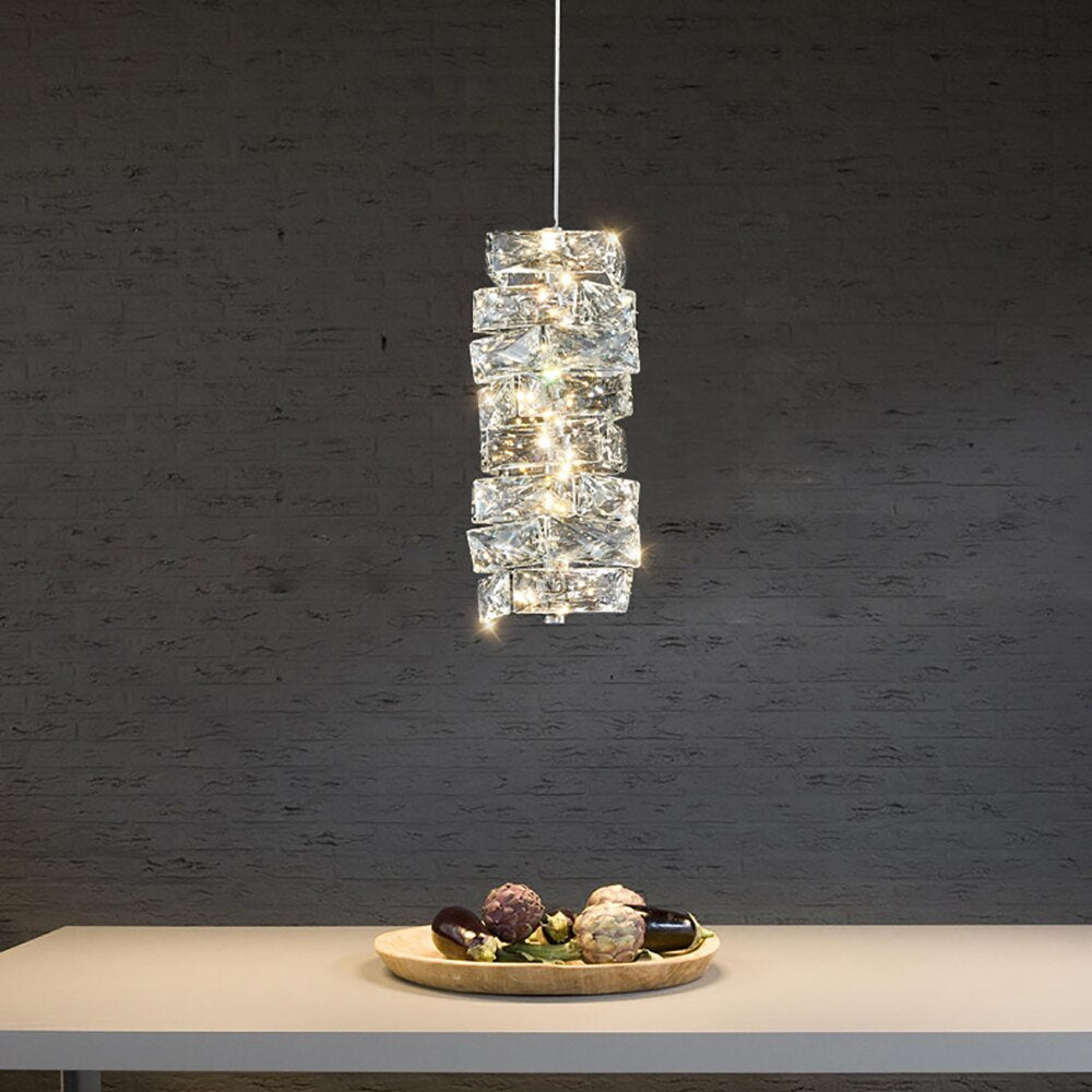 A Bacci Crystal Pendant Light Fixture made by Morsale.com hangs above a white table displaying a decorative tray with colorful stones, set against a dark textured wall.