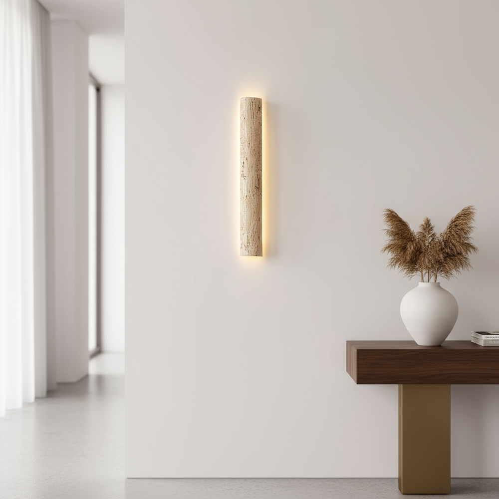 A modern interior features the Morsale Solterra Travertine Sconce, a wooden console with gold accent, and a white vase of pampas grass, all set against a white wall with sheer curtains.