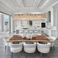 A modern dining room features a wooden table, eight white upholstered chairs, and three vases. Above, the Gio 47" Crystal Dining Room Chandelier by Morsale.com adds elegance. In the background: a white kitchen and large windows with water views.