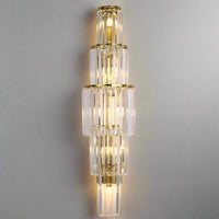 The Gio Crystal Wall Sconce by Morsale.com features tiered, gold-framed clear crystal rods in a symmetrical pattern. Dimmable LED bulbs emit a warm glow through the glass, enhancing any neutral wall with elegance.