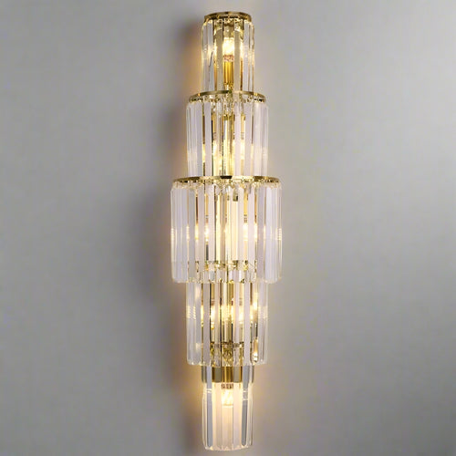 The Gio Crystal Wall Sconce by Morsale.com features tiered, gold-framed clear crystal rods in a symmetrical pattern. Dimmable LED bulbs emit a warm glow through the glass, enhancing any neutral wall with elegance.