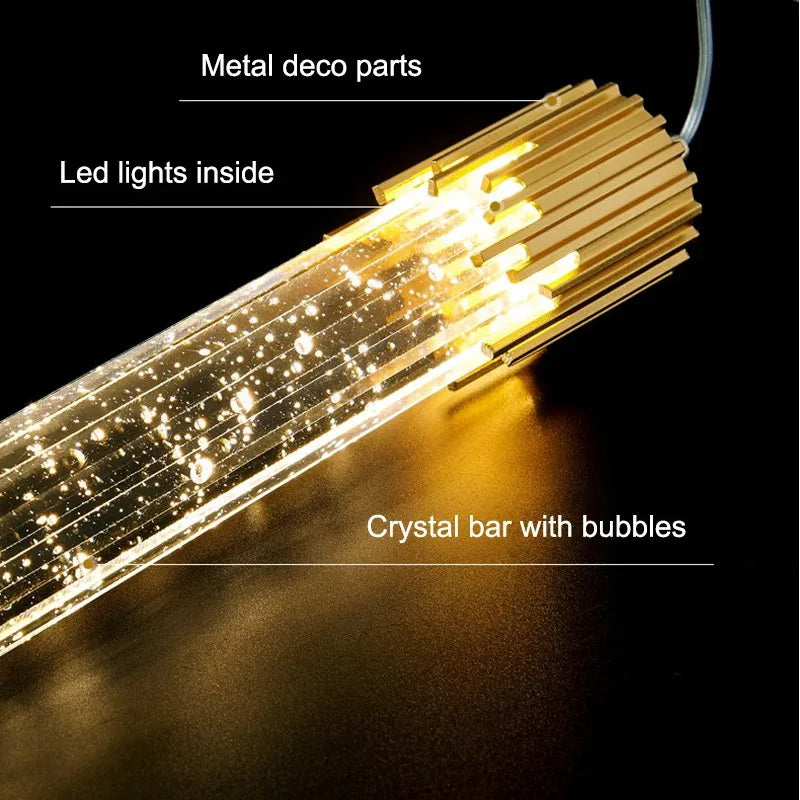 A close-up of the Modern Minimalist LED Pendant Light by Morsale.com is shown. It features polished gold decorative parts, dimmable LED lights inside, and a crystal bar with bubbles. The light is turned on, illuminating the bubbles within the crystal bar. Labels point to each component.