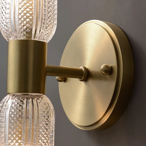 A close-up of the Palermo 12" Brass Wall Light Sconce by Morsale with intricate glass detailing. The stylish design features a circular brass plate mounted on a dark gray wall, with a straight arm holding the decorative glass elements that are subtly illuminated, providing warm lighting to the space.