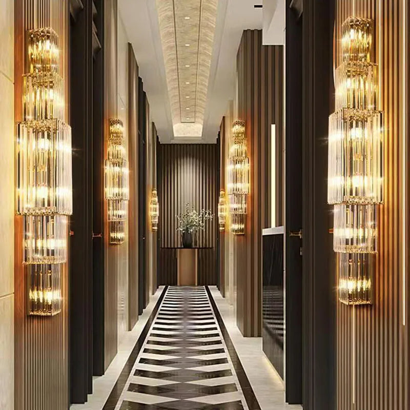 A luxurious hallway showcases elegant home decor, featuring Morsale.com Gio 32" Crystal Wall Sconces casting a warm glow on the marble-patterned floor. The dark wood walls complement a vase of white flowers at the corridor's end beneath a mirrored ceiling.