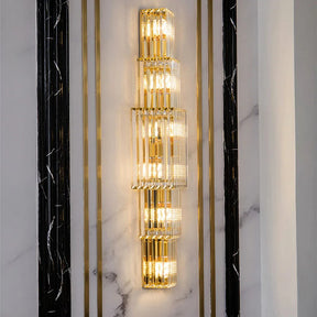 The Gio 32" Crystal Sconce by Morsale.com is a tall, elegant fixture featuring multiple rectangular, stacked glass panels that glow warmly against a white and black marbled wall, epitomizing luxury home decor.