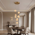 A modern dining room features a round wooden table, four beige chairs, and the Morsale.com Auris 9-Light Marble Chandelier with adjustable cords. Neutral tones, large curtained windows, and decorative accents complete the space.