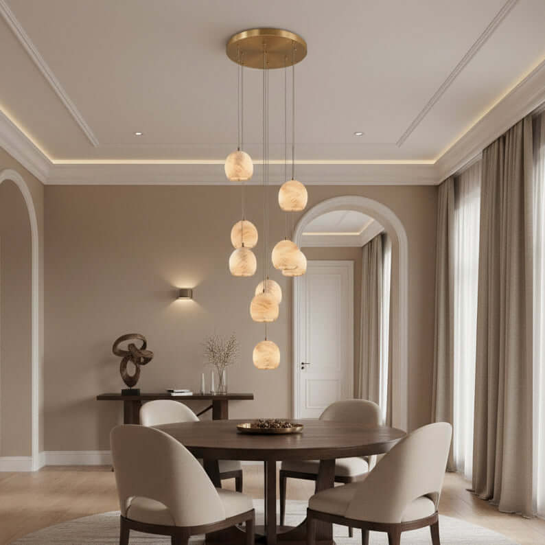 A modern dining room features a round wooden table, four beige chairs, and the Morsale.com Auris 9-Light Marble Chandelier with adjustable cords. Neutral tones, large curtained windows, and decorative accents complete the space.