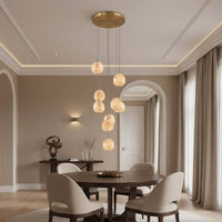 A modern dining room features a round wooden table, four beige chairs, and the Morsale.com Auris 9-Light Marble Chandelier with adjustable cords. Neutral tones, large curtained windows, and decorative accents complete the space.