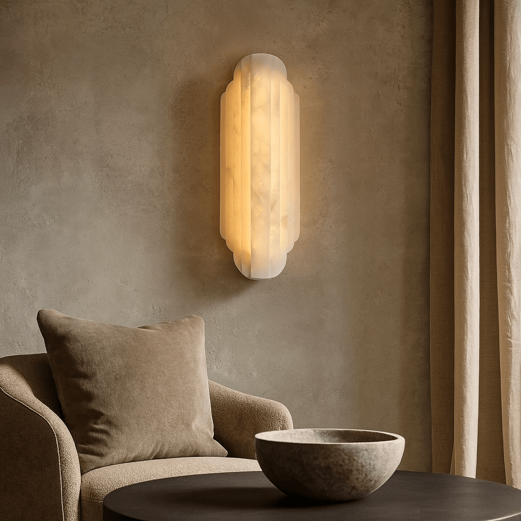 A soft beige lounge chair with a matching cushion sits by a round, dark coffee table. The Morsale Arina Marble Sconce glows warmly on a textured taupe wall, creating a cozy, minimalist ambiance.