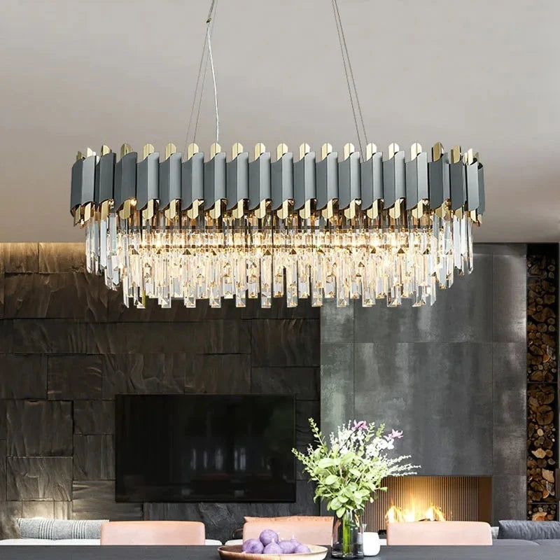 A modern dining room features a large, rectangular Gio 39" Crystal Dining Room Chandelier by Morsale.com with geometric design elements and dimmable LED lights. Below it is a gray dining table surrounded by leather chairs. On the table is a vase with flowers and a bowl of purple vegetables. In the background is a stone wall and a fireplace.