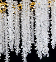 Close-up of crystal strands hanging from the Albero Crystal Foyer Chandelier by Morsale.com against a black background. The strands, composed of numerous clear, faceted crystals, sparkle with reflected light, creating a luxurious and elegant visual effect. The chandelier’s polished gold frame is partially visible.