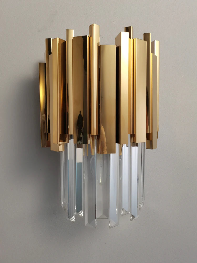 The Gio Crystal Wall Sconce by Morsale.com is a gold electroplated stainless steel wall-mounted light fixture featuring vertical rectangular elements and hanging crystal prisms. Illuminated by E14 LED bulbs, the fixture casts a warm glow. The logo "JASON" is in the top-left corner.