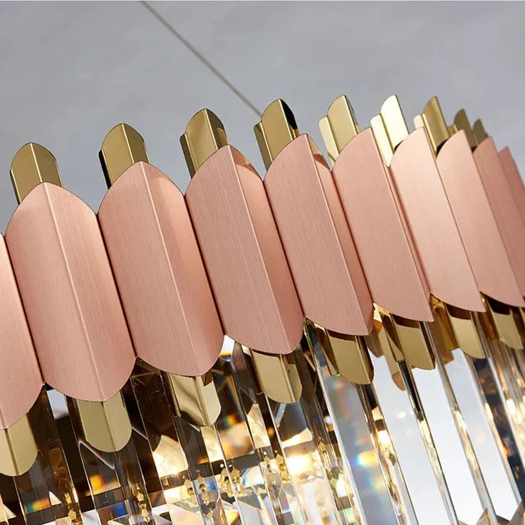 Close-up of the Morsale.com Gio 39" Rose Gold Dining Room Chandelier featuring a combination of stainless steel and iron materials. The design includes pink and gold metallic elements arranged in alternating patterns, with hanging crystal prisms that reflect light. Text at the bottom reads "Stainless steel + iron lamp body.