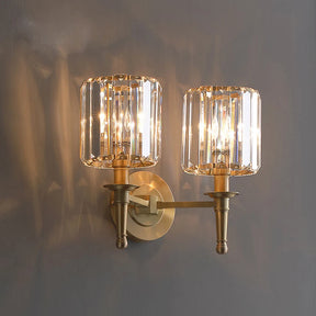 The Morsale Gio Crystal Sconce Wall Light is a wall-mounted, double-arm brass fixture with two cylindrical glass shades and LED bulbs that cast warm light and subtle reflections on a gray wall, blending elegant classic styling with modern flair.