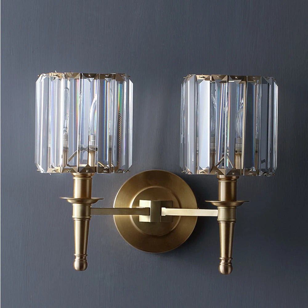 The Gio Crystal Sconce Wall Light by Morsale features a modern brass design with two cylindrical clear crystal lampshades, energy-efficient LED bulbs, a circular base, and elegant straight arms that illuminate beautifully on a dark gray wall.