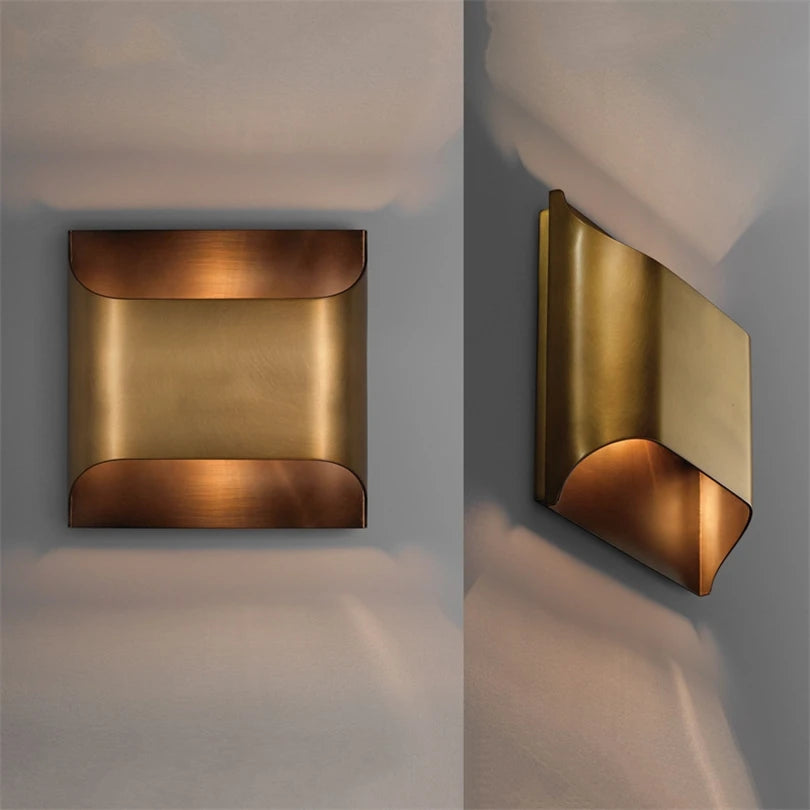 The Rame Modern Copper Wall Lights by Morsale are wall-mounted fixtures with a sleek geometric design, made from polished brass. With a square front and an angled side view, these lamps cast a warm glow using LED bulbs against a gray backdrop.