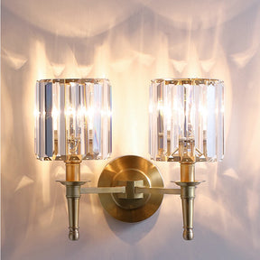 The Gio Crystal Sconce Wall Light by Morsale is a modern dual wall sconce with a round gold base and two cylindrical crystal shades, each holding an LED bulb. It casts a warm glow on the wall.