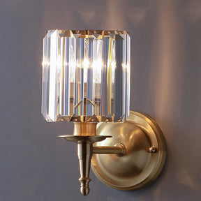 The Morsale Gio Crystal Sconce Wall Light combines modern style with a classic touch, featuring a brass mount and a clear geometric crystal lampshade. Equipped with LED bulbs, it illuminates a smooth gray wall with warm, elegant light.