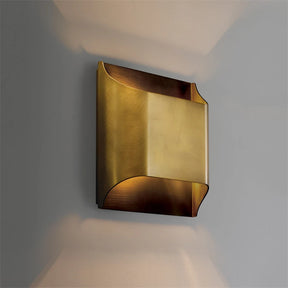 The Rame Modern Copper Wall Lights by Morsale feature a rectangular brass shade, beautifully mounted on a gray wall. These lights emit a warm, ambient glow that evokes the charm of vintage wall lamps, tastefully accentuating the fixture's curved edges.