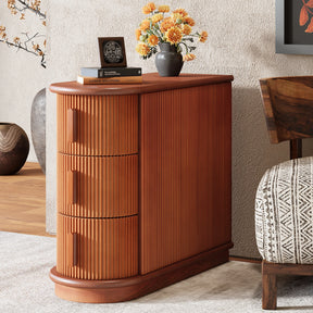The Morsale Galway Solid Wood End Table with three drawers displays a vase of orange flowers, a small book, and a decorative item on its top. It rests on a light-colored rug next to a patterned cushion, with an elegant wooden bowl visible underneath.