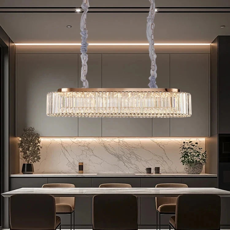 A modern dining room showcases a long rectangular table surrounded by chairs, with the elegant Gio Crystal Dining Room Chandelier by Morsale hanging above. The space features a marble backsplash, minimalistic decor, and soft, ambient lighting.